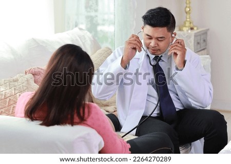 Happy smiling young asian pregnant female and male doctor sitting on sofa in living room at home with private treatment and consultant. Friendly doctor listening to baby’s heartbeat with stethoscope.