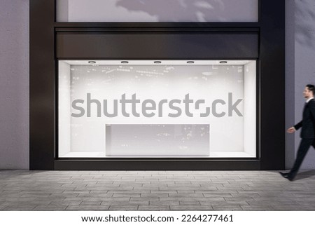 Man walking by evening empty shop window with podium inside for your product presentation on glowing white background in modern building area outdoors, mock up Royalty-Free Stock Photo #2264277461