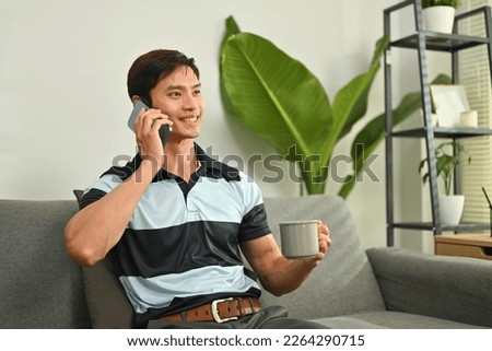 Handsome millennial man drinking coffee having pleasant phone conversation on couch at home