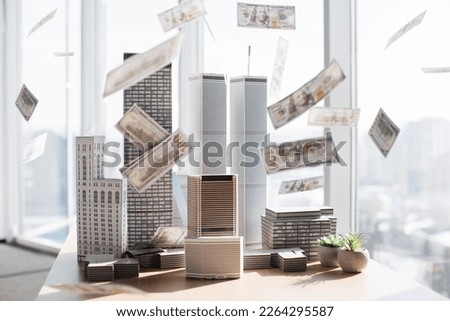 Table with paper dollars hundred banknotes falling on architectural scale cardboard model of building complex in empty modern architect office.