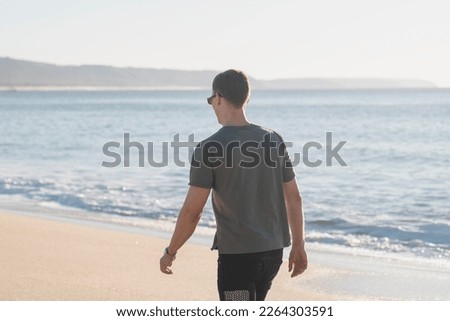 Young healthy beautiful white male on vacation walking on the beach in sun with waves in the ocean in the background