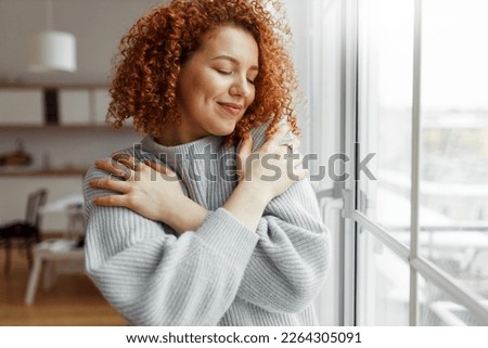 Horizontal image of pretty redhead female with closed eyes wearing rings on fingers, hugging herself, touching new soft sweater, enjoying comfort of fabric, standing next to panoramic window at home Royalty-Free Stock Photo #2264305091