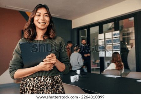 Cheerful young businesswoman smiling at the camera while holding a digital tablet. Happy young businesswoman standing in a boardroom with her colleagues in the background. Royalty-Free Stock Photo #2264310899