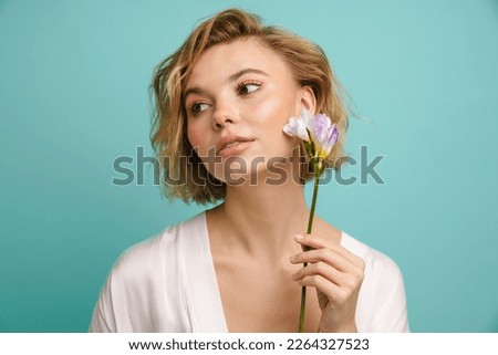 Young beautiful blonde woman posing with freesia flower at her face isolated over blue studio background
