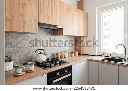 Interior of small kitchen with sink near window and wooden cabinets in kitchen furniture. Stove with gas and kettle. Modern pattern tiles on the wall. Royalty-Free Stock Photo #2264333491