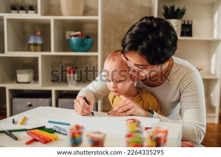 Family of two improving fine motor skill trough the games while drawing and coloring with crayons. Toddler boy playing with his mother or aunt. Funny educational games for children. Copy space. Royalty-Free Stock Photo #2264335295