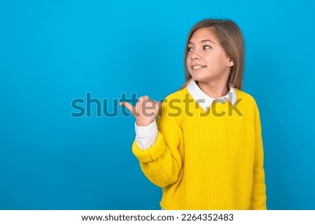 Charming beautiful caucasian teen girl wearing yellow sweater over blue wall looking at copy space having advertisements