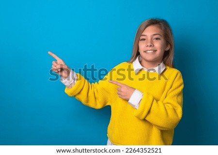 beautiful caucasian teen girl wearing yellow sweater over blue wall indicating finger empty space showing best low prices, looking at the camera