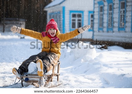 A little funny boy rides on a wooden sled in the village on a winter day.