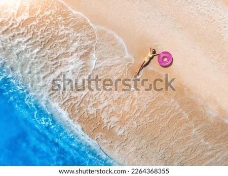 Aerial view of the lying beautiful young woman with pink swim ring on the sandy beach near sea with waves at sunny day. Summer in Lefkada island, Greece. Drone view of slim girl, clear blue water