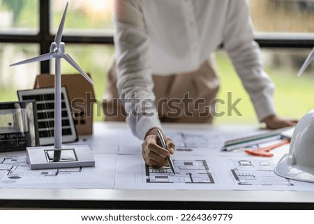 Female engineer points to blueprints for house hoist and wind turbine next to concept of building house with renewable energy installation with wind turbine and solar cells Royalty-Free Stock Photo #2264369779