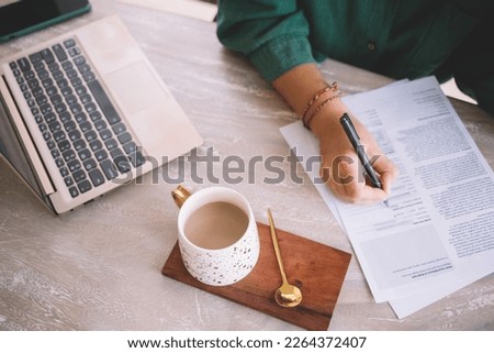 Cropped view of unrecognizable business woman with milk tea and modern laptop computer analyzing documents during working process in cafe interior, female entrepreneur checking report information