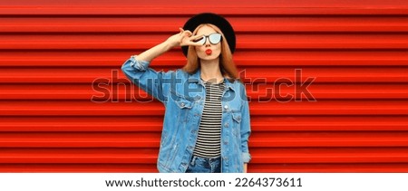 Portrait of stylish young woman model blowing her lips sending sweet air kiss wearing jean jacket, black round hat on red background Royalty-Free Stock Photo #2264373611