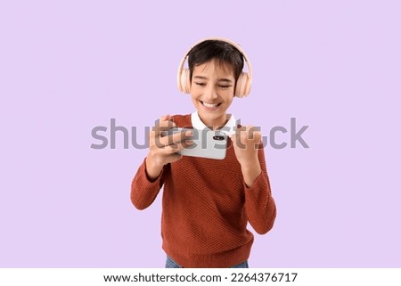 Happy little boy in headphones with mobile phone on lilac background