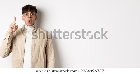 Portrait of young man student pitching an idea, raising finger up and say suggestion, have a plan, standing on white background in glasses and casual clothes.