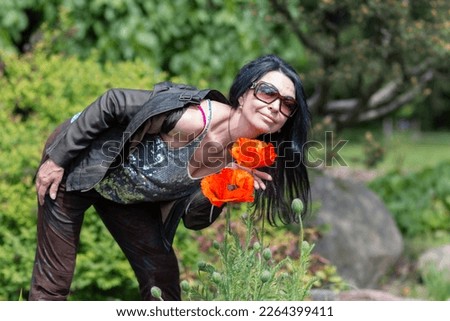 A happy girl with long black hair in glasses sniffing the aroma of large red poppy petals in the garden. Fragrant poppy flowers. Blurred background. Selective focus.