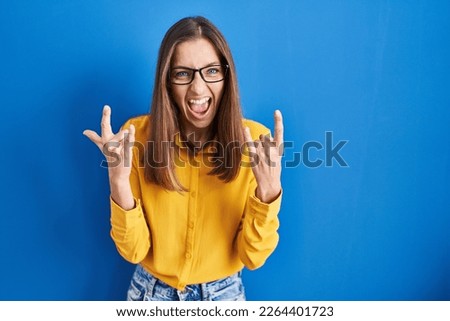 Young woman wearing glasses standing over blue background shouting with crazy expression doing rock symbol with hands up. music star. heavy concept. 