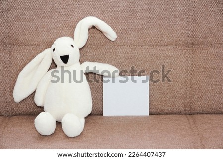 Children's toy white fluffy plush bunny holding a greeting card on a brown background