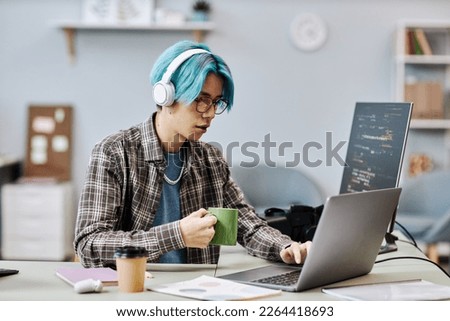 Portrait of young man with blue hair using computer in office and wearing headphones while writing code Royalty-Free Stock Photo #2264418693