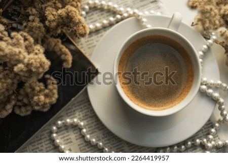 A white cup of coffee on a saucer, with a string of pearls, dried flowers, a magazine and a newspaper. Aesthetic picture, elegant background photo.