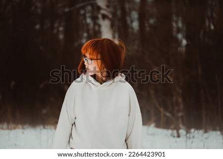 Portrait of a beautiful ginger Asian woman in a warm white hoodie against the background of a dark park on a winter day. Lifestyle and nature walk.