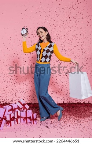 joyful and fashionable woman posing with alarm clock and shopping bags near confetti and gift boxes on pink background