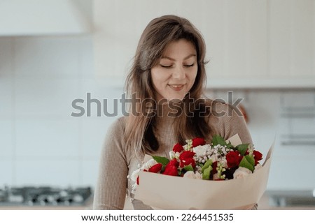 Beautiful young woman wearing casual fashion clothes holds a bouquet of flowers and looking at camera while standing on modern kitchen. Cozy atmosphere.Women day or special date concept. 