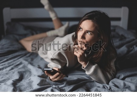 Young happy smile woman day dreaming think looking up hold cell phone on bed in bedroom, at home, girl lying type message, look at window, flirting. Royalty-Free Stock Photo #2264451845