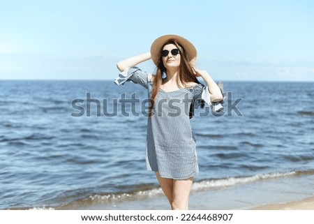 Happy young brunette woman standing on the ocean beach while smiling, and wearing fashion hat, sunglasses. The enjoying vacation concept.