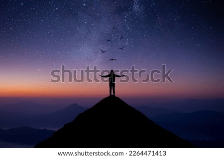 Silhouette of young man standing alone on top of mountain and raise both arms praying and free bird enjoying nature on beautiful night sky, star, milky way background. Demonstrates hope and freedom.