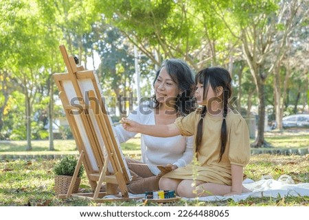 Loving grandmother and little granddaughter enjoy painting picture together in the park surrounded by green trees.