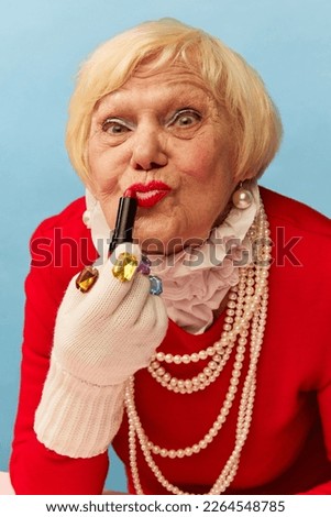 Applying lipstick. Beautiful old woman, grandmother in stylish red dress and pearl necklace posing over blue studio background. Concept of age, fashion, lifestyle, emotions, facial expression