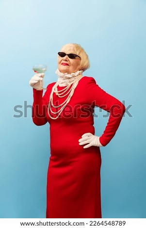 Beautiful old woman, grandmother in stylish red dress and pearl necklace posing, drinking champagne over blue studio background. Concept of age, fashion, lifestyle, emotions, facial expression