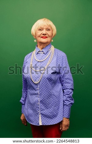 Beautiful old woman, grandmother in purple shirt and pearl necklace posing, smiling over green studio background. Extravagant look. Concept of age, fashion, lifestyle, emotions, feelings