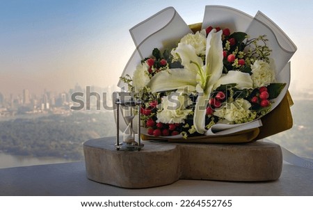 Hourglass or Sandglass is next to Bouquet of flowers placed on the timber outside the balcony with the city view background. Symbol of Romantic forever love, Free space for text.