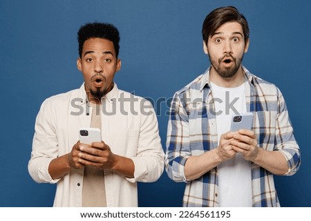 Young two friends surprised happy men 20s wear white casual shirts together hold in hand use mobile cell phone surfing internet isolated plain dark royal navy blue background. People lifestyle concept
