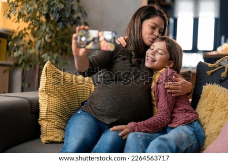 Beautiful caring pregnant single mother kissing her lovely adorable little girl, holding smartphone, capturing precious moments, taking selfie or recording vlog for social media. Selective focus.