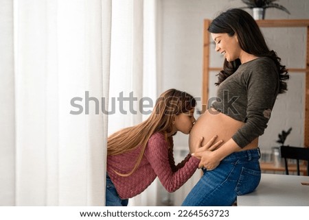 Lovely little girl kissing belly of a pregnant mother. Mother in advanced stage of pregnancy with her adorable girl joyfully bonding at home. Caring little girl giving kisses to baby in mom's tummy.