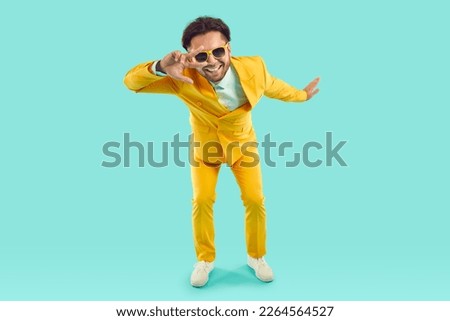 Cool funny man showing v-sign and looking at you through it on light blue background in studio. Stylish man in bright yellow suit and sunglasses smiling making v-sign near eyes. Full length.