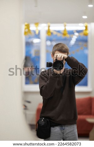 a male photographer takes a selfie on a camera in the mirror, he is dressed in jeans and a brown sweatshirt. photographer taking a picture in a mirror with a mirrorless camera