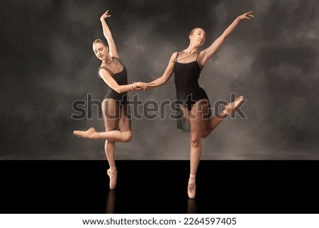 Two young women ballet dancers in black leotards and dark veil coverlets, dancing on a black floor in a Connecticut studio, against a gray background.