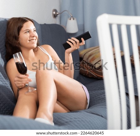 Happy young woman watching tv relaxing on sofa with glass of wine at condo apartment