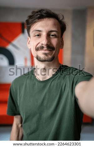 One man young adult caucasian male standing at home with brown hair mustaches and beard looking to the camera happy smile confident real people green t shirt UGC selfie user generated content Royalty-Free Stock Photo #2264623301