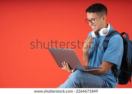 Young man with glasses and white headphones on his neck, sitting cross-legged being happy and laughing while making a video call on a laptop, on a red background.