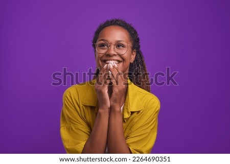 Young cheerful ethnic African American woman in glasses and casual clothes laugh covering mouth with hands watching comedy series or evening TV show stands in purple studio. Humor, joke Royalty-Free Stock Photo #2264690351