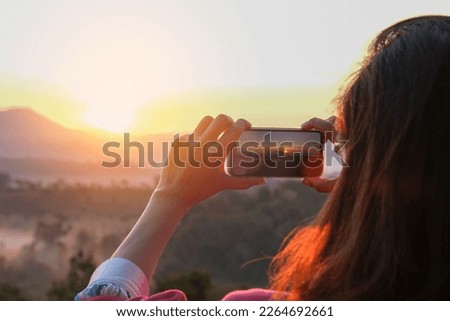 A woman uses her smartphone to take pictures of the sunrise in a valley with abundant nature tourism and technology. Royalty-Free Stock Photo #2264692661