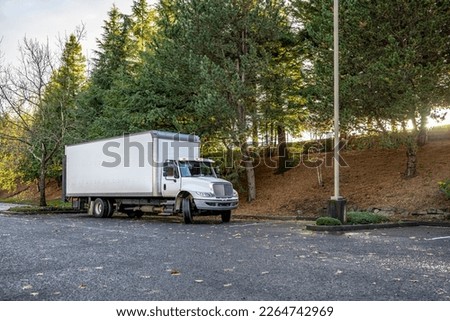 White middle rig semi truck with day cab for local deliveries with long box semi trailer standing for rest on the street parking lot at industrial area with tree alley waiting for the next load Royalty-Free Stock Photo #2264742969