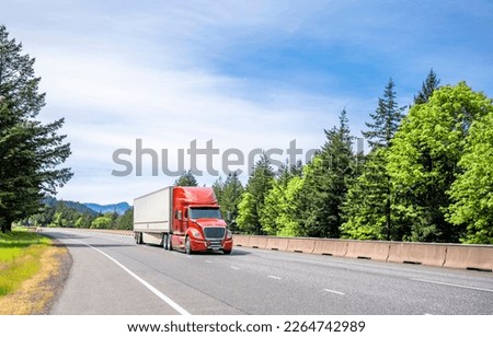 Red big rig long haul industrial semi truck tractor transporting cargo in dry van semi trailer running on curving highway road with protective fence and green mountain forest in Columbia Gorge Royalty-Free Stock Photo #2264742989