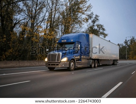 Industrial powerful blue big rig semi truck with turned on headlights transporting commercial cargo in dry van semi trailer driving on the twilight evening highway road for timely delivery Royalty-Free Stock Photo #2264743003