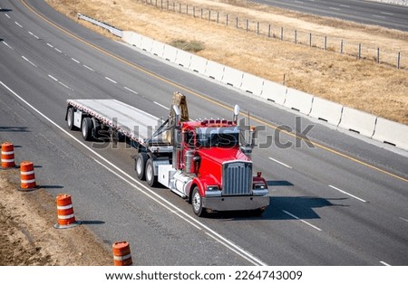Classic bonnet red semi truck with additional side lights and tall chrome exhaust pipes transporting cargo on flat bed semi trailer running on the highway road to warehouse for delivery unload Royalty-Free Stock Photo #2264743009